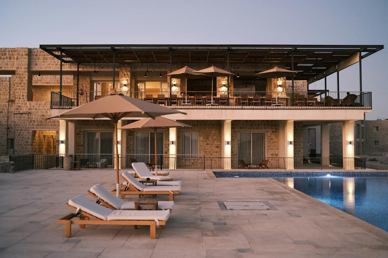 A stone resort terrace with sun loungers beside a lit pool at dusk
