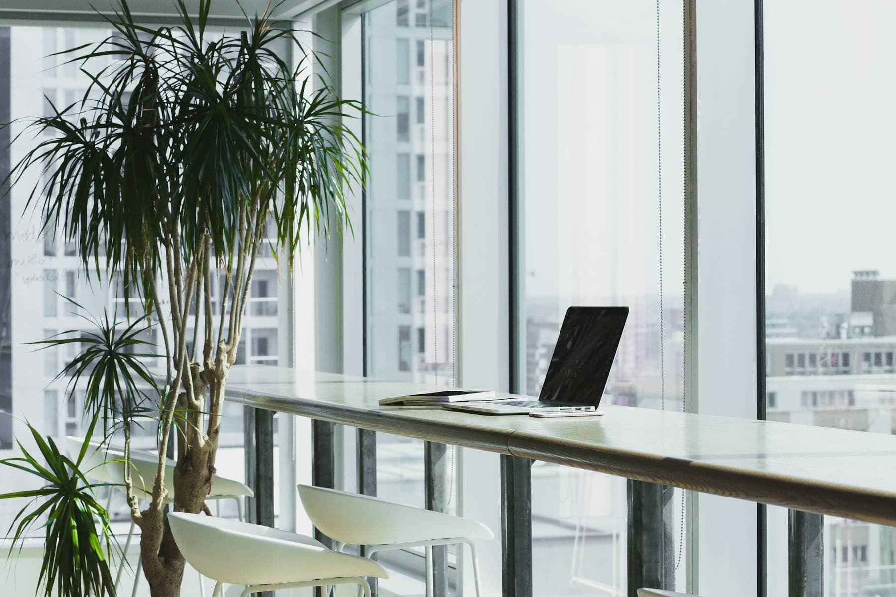 A bright office with a laptop on a long desk beside floor-to-ceiling windows and city towers